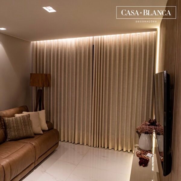 Uma sala de estar moderna com um sofá de couro marrom, almofadas decorativas, uma luminária de piso de madeira, cortinas bege, uma TV de parede e uma mesa de console com vasos e flores. O logotipo da Casa Blanca Decorações está no canto superior direito.