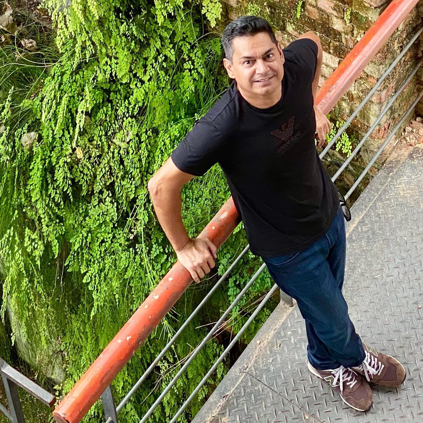 Um homem de camiseta preta e calça jeans azul está em uma passarela de metal, segurando uma grade vermelha. Ele está sorrindo e olhando para a câmera. Uma exuberante folhagem verde cresce na parede atrás dele.