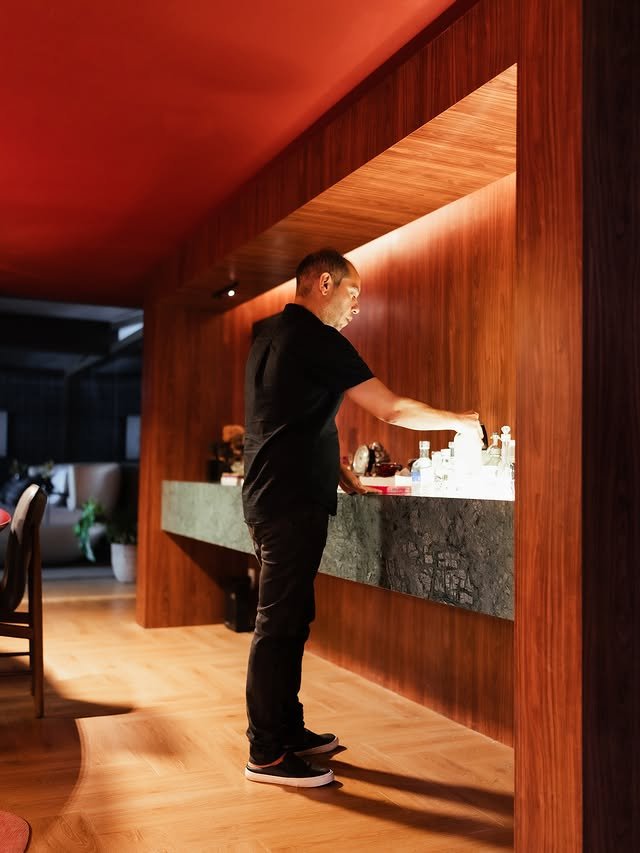 Um homem em uma roupa preta está em um moderno balcão de bar de madeira com garrafas e copos, sob uma iluminação quente em um interior elegante.