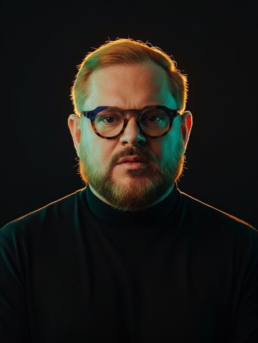 Um homem com cabelo castanho-claro curto, barba e óculos usa uma gola alta preta, olhando diretamente para a câmera. O fundo é escuro e a iluminação verde-amarelada destaca a lateral de seu rosto.